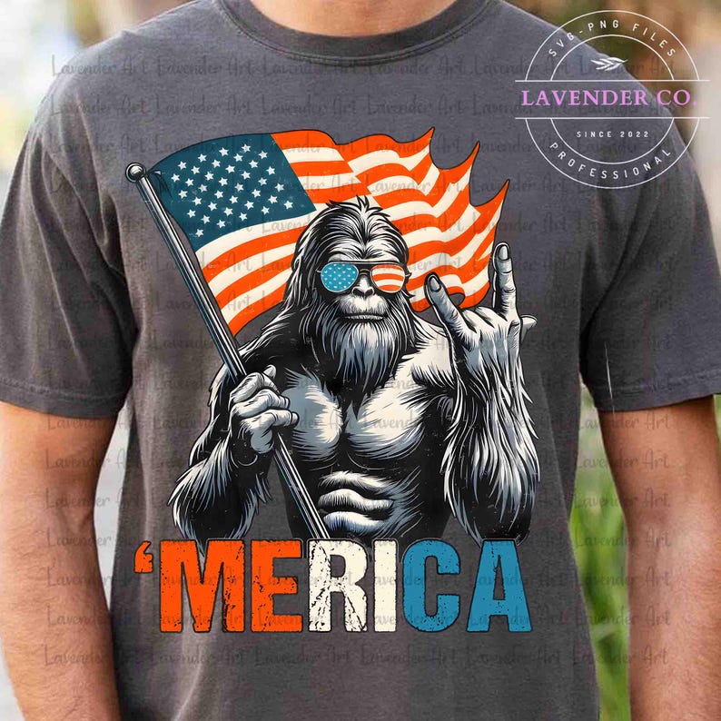 Puede incluir: Camiseta gris oscuro con un dise&ntilde;o de Bigfoot con gafas de sol con la bandera estadounidense, sosteniendo una bandera estadounidense y haciendo el gesto de rock. La palabra "'MERICA" est&aacute; impresa en rojo, blanco y azul. La camiseta tiene el texto "LAVENDER CO" y "SINCE 2022".