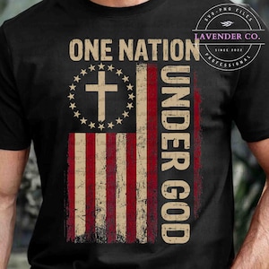 May include: Black t-shirt featuring a distressed American flag design with a cross and the text "ONE NATION UNDER GOD." The flag has red and white stripes, with a circle of stars around the cross.