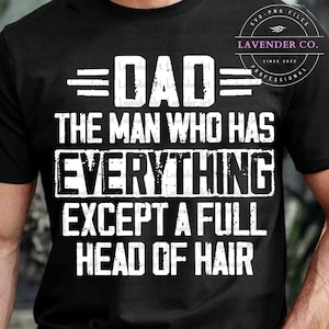 以下が含まれることがあります： 「DAD THE MAN WHO HAS EVERYTHING EXCEPT A FULL HEAD OF HAIR」と白い文字で書かれた黒いTシャツ。シャツにはLavender Co.のロゴも表示されています。
