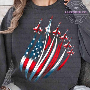 May include: Dark gray sweatshirt featuring a patriotic design. The graphic depicts jets streaking across the sky, leaving trails of red, white, and blue, forming the American flag. The text "Lavender Co." is in a circle.