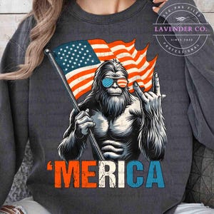 Puede incluir: Sudadera gris oscuro con un dise&ntilde;o de Sasquatch con gafas de sol con el dise&ntilde;o de la bandera estadounidense. El Sasquatch sostiene una bandera y hace un gesto de rock and roll con la mano. La palabra "'MERICA" est&aacute; impresa en rojo, blanco y azul.