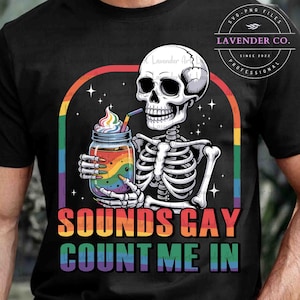 May include: Black t-shirt featuring a skeleton drinking from a rainbow-colored jar with whipped cream. The text "SOUNDS GAY COUNT ME IN" is displayed in rainbow colors. The shirt also has the text "LAVENDER CO." and "SVG-PNG FILES".