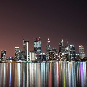 Puede incluir: Una vista panorámica nocturna de un horizonte urbano con edificios altos reflejados en el agua. Los edificios están iluminados, creando un reflejo brillante y colorido en el agua.