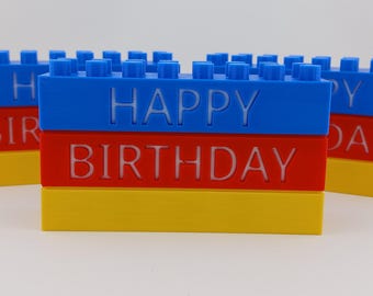 May include: Three stacks of colorful plastic building blocks spelling out "Happy Birthday". Each stack is made of three blocks: blue, red, and yellow.