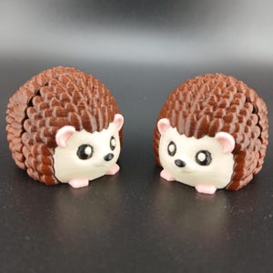 May include: Two hedgehog figurines. Each has a brown, textured body, white face, pink ears and feet, and black eyes. The figurines are displayed on a dark surface.