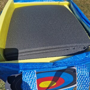 May include: A blue and yellow archery target bag with a black foam target inside. The bag has two handles and a zipper closure.