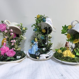 May include: Three decorative teacup arrangements featuring miniature princess figurines in pink, blue, and yellow dresses. Each teacup is tilted, spilling artificial greenery, moss, and colorful flowers onto matching saucers, creating a garden scene.