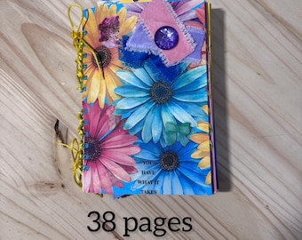 May include: A handmade journal with a floral design featuring pink, yellow, and blue flowers. The journal measures 9 x 5 x 3 inches and has 38 pages. Embellishments include a purple jewel and ribbon.