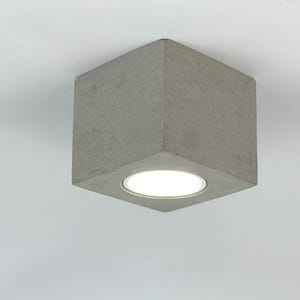 May include: A square, concrete ceiling light fixture with a round, white light in the center.