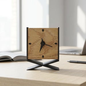 May include: A square wooden desk clock with a light brown face and black hands and frame. The clock has a unique black base. A laptop, pen, and open book are also on the desk.