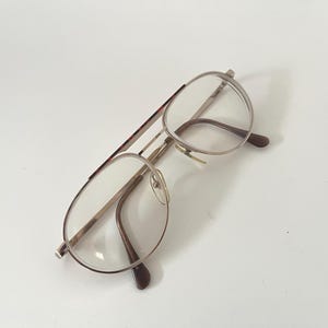 Könnte beinhalten: Eine Vintage-Pilotenbrille mit einem goldfarbenen Metallrahmen und klaren Gläsern. Die Bügel haben ein braunes Schildpattmuster. Die Brille liegt auf einer weißen Oberfläche.