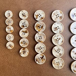 May include: A collection of various-sized buttons with a marbled, cream and brown appearance. The buttons have four holes and are arranged in rows on a brown surface. The buttons are likely for sewing or crafts.