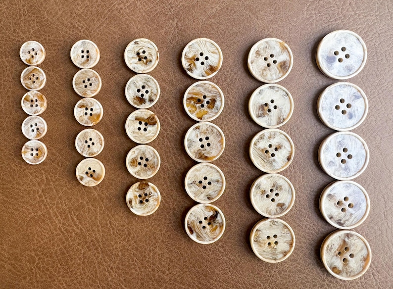 May include: A collection of various-sized buttons with a marbled, cream and brown appearance. The buttons have four holes and are arranged in rows on a brown surface. The buttons are likely for sewing or crafts.