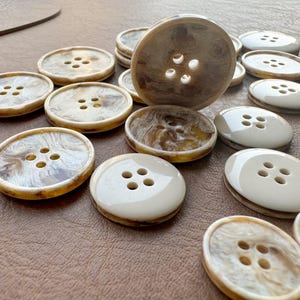 May include: A collection of assorted buttons in shades of cream, brown, and white. The buttons have four holes and a glossy finish. Some buttons have a marbled effect. The buttons are arranged on a brown surface.