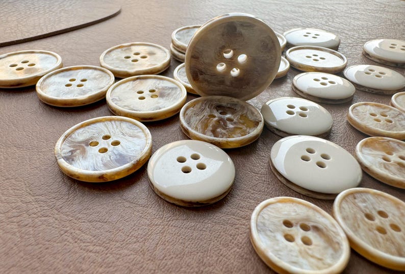 May include: A collection of assorted buttons in shades of cream, brown, and white. The buttons have four holes and a glossy finish. Some buttons have a marbled effect. The buttons are arranged on a brown surface.