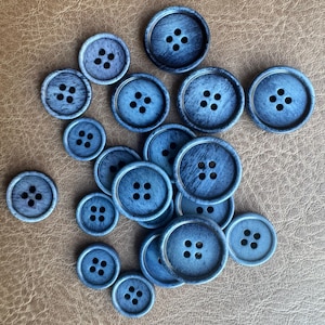 Op de afbeelding: Een verzameling ronde, blauwe knopen van verschillende grootte, elk met vier gaten. De knopen hebben een verweerde, getextureerde uitstraling en liggen op een bruin oppervlak. Ideaal voor naai- en knutselprojecten.