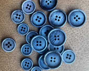 May include: A collection of various sized, round, blue buttons with four holes each. The buttons have a distressed, textured appearance and are scattered on a brown surface. Ideal for sewing and craft projects.