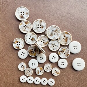 May include: A collection of round buttons in various sizes, scattered on a brown surface. Some buttons are solid white with four holes, while others have a marbled brown and white pattern. The buttons are made of a hard, smooth material.
