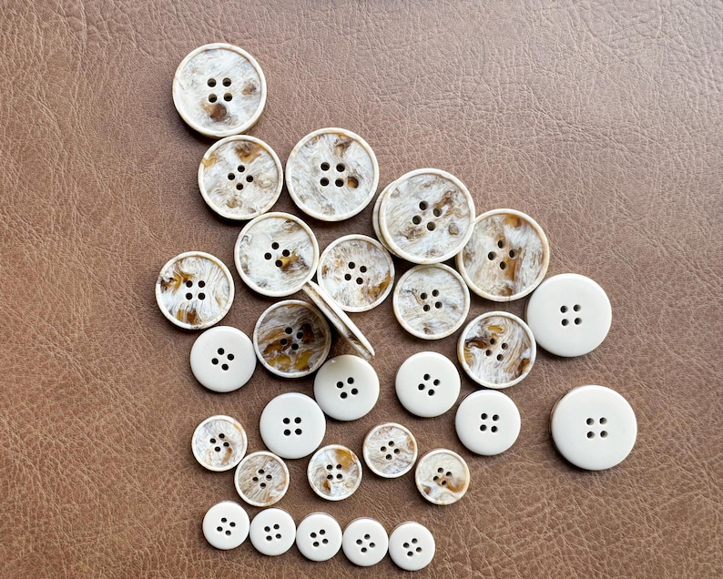 May include: A collection of round buttons in various sizes, scattered on a brown surface. Some buttons are solid white with four holes, while others have a marbled brown and white pattern. The buttons are made of a hard, smooth material.