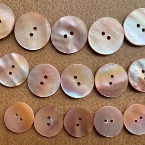 Peut inclure: Une collection de boutons ronds et irisés, chacun avec deux ou trois trous. Les boutons varient en couleur, allant du beige clair à une teinte pêche chaude. Les boutons sont disposés sur une surface brune.