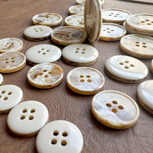 May include: A collection of various sized buttons with four holes. The buttons have a cream-colored outer ring and a marbled, tan-colored center. Some buttons are oval, while others are round. The buttons are scattered on a brown surface.
