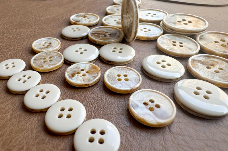 May include: A collection of various sized buttons with four holes. The buttons have a cream-colored outer ring and a marbled, tan-colored center. Some buttons are oval, while others are round. The buttons are scattered on a brown surface.