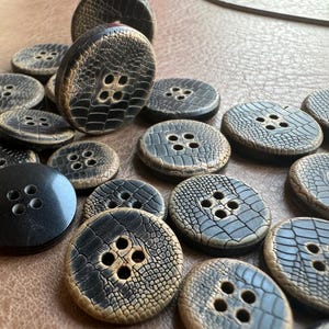 Puede incluir: Una colección de botones de varios tamaños y colores oscuros, con cuatro agujeros. Los botones tienen un patrón texturizado, similar a la piel de reptil, en tonos marrones y grises. Algunos botones son negros sólidos. Los botones están esparcidos sobre una superficie marrón.