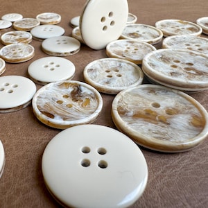 May include: A collection of ivory and marbled buttons with four holes each. The buttons vary in size and have a glossy finish. Some buttons have a solid ivory color, while others feature a brown and cream marbled pattern.