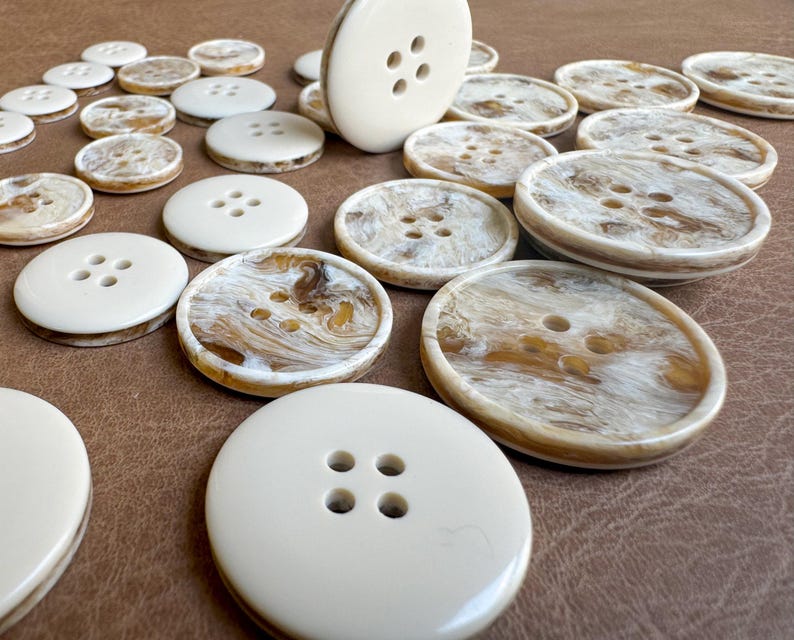 May include: A collection of ivory and marbled buttons with four holes each. The buttons vary in size and have a glossy finish. Some buttons have a solid ivory color, while others feature a brown and cream marbled pattern.