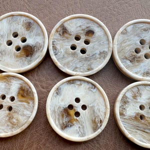 Peut inclure: Six gros boutons ronds avec un motif marbré blanc cassé et marron. Chaque bouton comporte quatre trous pour la couture. Les boutons sont disposés sur une surface marron.