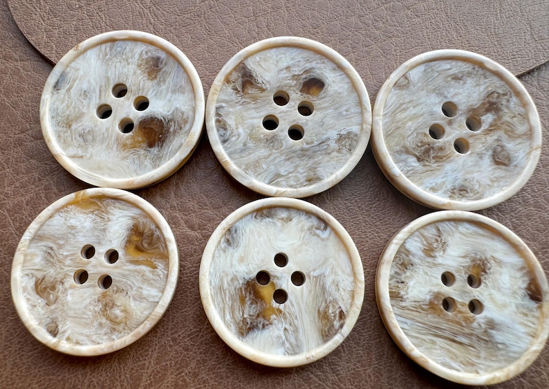 May include: Six large, round buttons with a marbled, off-white and brown pattern. Each button has four holes for sewing. The buttons are arranged on a brown surface.