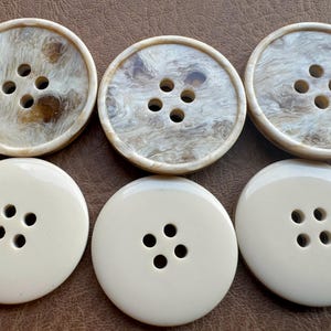 May include: Six large, round buttons. Three buttons have a marbled, tan and cream pattern, each with four holes. The other three are solid cream-colored with four holes. All buttons are on a brown surface.