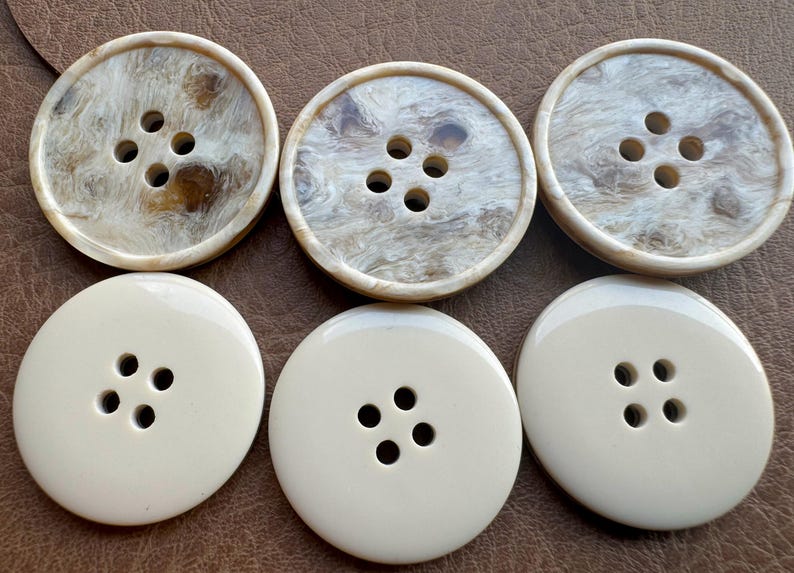 May include: Six large, round buttons. Three buttons have a marbled, tan and cream pattern, each with four holes. The other three are solid cream-colored with four holes. All buttons are on a brown surface.