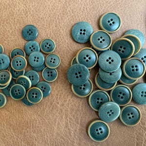 May include: A collection of teal-colored buttons with four holes each, arranged in two groups. The buttons have a light tan rim and are scattered on a brown surface. These buttons are suitable for sewing and craft projects.