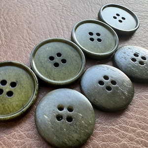 May include: A collection of seven olive green buttons with four holes each. The buttons are round and have a slightly textured surface. The buttons are arranged on a brown leather-like surface.