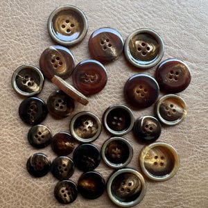May include: A collection of round, four-hole buttons in various shades of brown, ranging from dark espresso to light amber. The buttons feature smooth, glossy surfaces, with some displaying a subtle marbled pattern and others having a ridged edge.