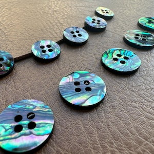 Può includere: Una collezione di bottoni iridescenti in conchiglia di abalone di varie dimensioni. I bottoni hanno una lucentezza perlescente con sfumature di blu, verde e viola. Ogni bottone presenta quattro fori per cucire ed è disposto su una superficie marrone.