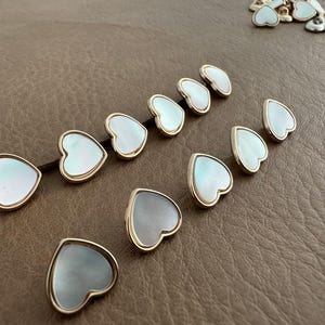 May include: A collection of heart-shaped jewelry components with iridescent mother-of-pearl inlays and gold-colored metal edges. The components are arranged on a brown surface, showcasing their delicate design and craftsmanship.