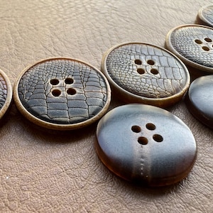 Pode incluir: Uma coleção de sete botões redondos com quatro furos cada. Os botões apresentam um padrão texturizado castanho, semelhante à pele de crocodilo, e um acabamento liso castanho escuro. Estão dispostos numa superfície castanha.