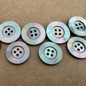 Può includere: Una collezione di dieci bottoni iridescenti, ciascuno con quattro fori. I bottoni rotondi hanno una finitura madreperlacea, che mostra sfumature di blu, verde e viola. Sono disposti su una superficie marrone.