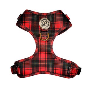 May include: Red and black tartan dog harness with a gold-coloured metal ring for leash attachment. The harness features a round patch with the text "Hendricks and Maple". The harness is designed to fit around the dog's chest and shoulders.