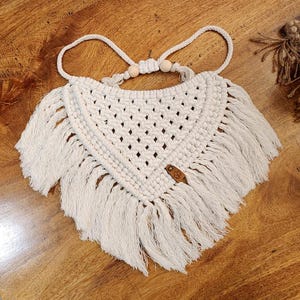 May include: An off-white macrame bandana featuring a triangular woven pattern with intricate knotting, long fringe edges, two light wooden beads on the adjustable cord, and a small brown leather-like tag with a butterfly logo embossed on it.