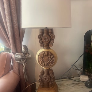 Puede incluir: Una lámpara de mesa decorativa con una base de madera tallada beige y una pantalla blanca. La base presenta intrincados diseños circulares y florales. La lámpara está colocada sobre una superficie con vetas de mármol.