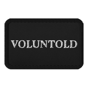 Op de afbeelding: Zwart rechthoekig embleem met het woord "VOLUNTOLD" in witte, hoofdletters geborduurd. De patch heeft een gestikte rand.