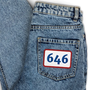 Puede incluir: Primer plano de unos vaqueros de mezclilla azul claro. El bolsillo trasero presenta un parche blanco con el número "646" en azul, enmarcado por un borde rojo. Los vaqueros están doblados, mostrando la textura y el lavado de la mezclilla.
