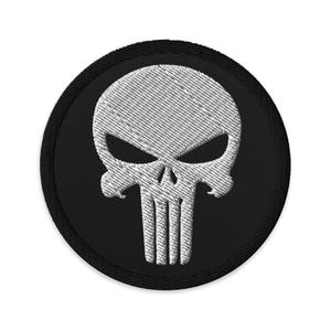 May include: Embroidered patch featuring a white skull design on a black circular background. The skull has detailed eye sockets, a nose cavity, and vertical lines for teeth. The patch has a black border.
