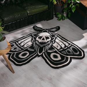 May include: A black, white, and grey moth-shaped rug with a skull design. The rug has a detailed wing pattern and a skull in the centre. It is placed on a light-coloured wooden floor.