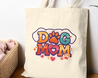 May include: Cream-colored tote bag with the words "DOG MOM" in colorful, retro-style lettering. The design includes a paw print, hearts, and a cloud-like shape. The bag has long handles and is made of a sturdy fabric.