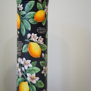 May include: A tall, cylindrical black container decorated with a lemon and floral design. The design features bright yellow lemons, white flowers, and green leaves. The container has a textured surface and a decorative top.