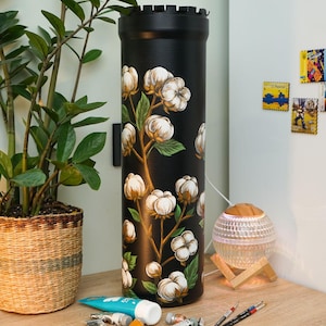 May include: A black cylindrical lamp with a floral cotton design. The lamp is decorated with white cotton flowers and green leaves. Paint tubes and brushes are on the wooden surface next to the lamp.